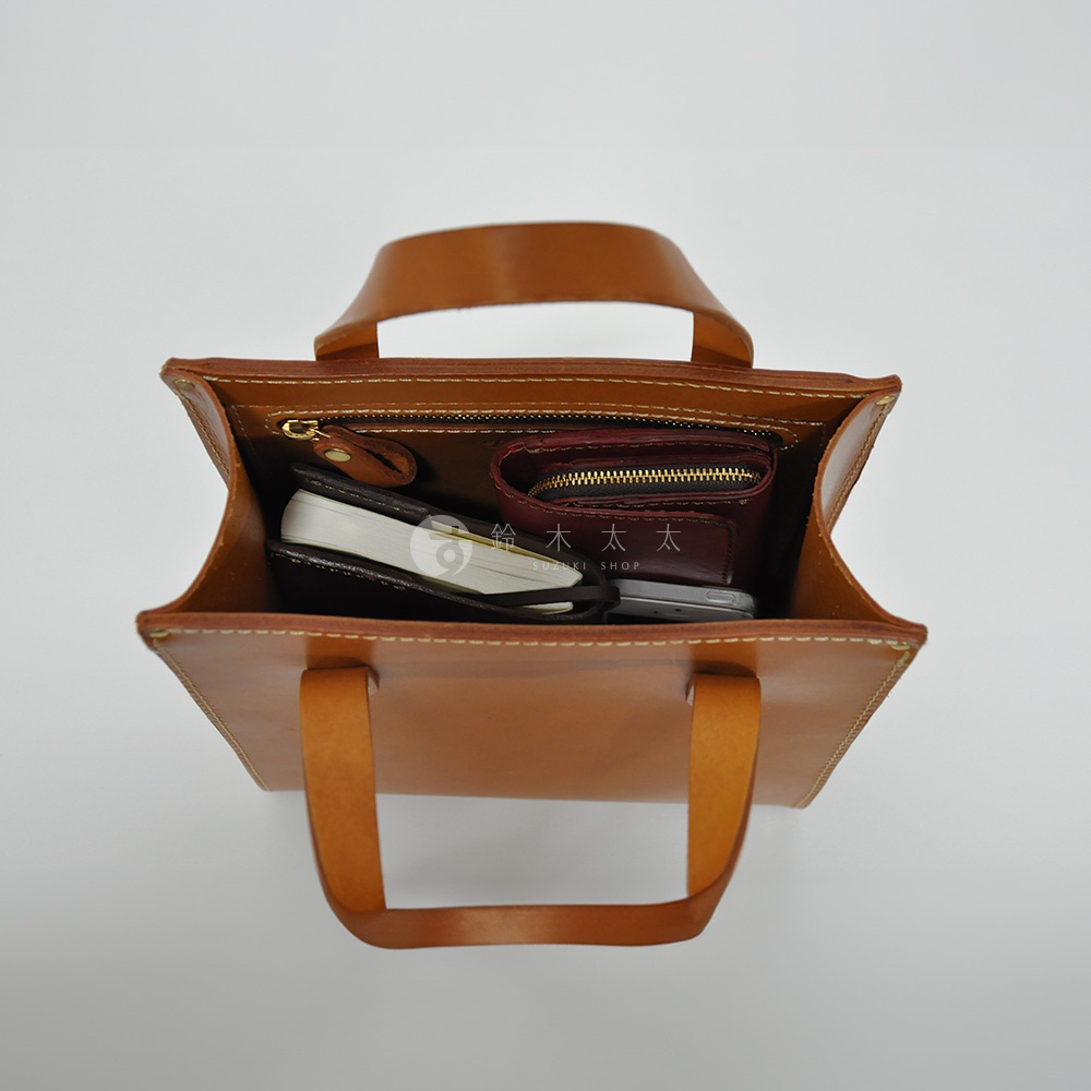 皮革直式托特包，顏色為淺棕色，包內裝有手機、筆記本和一個酒紅色拉鍊皮夾，整體以精緻縫線完成。