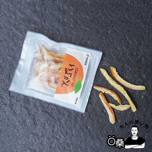 密封夾鏈袋裝著金黃色澤的柑橘果皮乾，部分散落在黑色紋理背景上。包裝上有日文標示與橘色漸層的柑橘圖案。