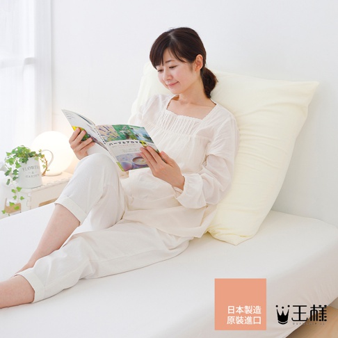 一位女性穿著白色居家服，坐在床上，抱著一個淺黃色的巨大枕頭，並閱讀一本雜誌。枕頭質地柔軟，看起來很舒適。