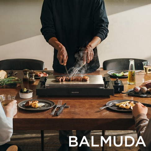 黑色極致電烤盤，配有銀色金屬烤盤，上面有切好的牛排正在烹飪，冒著煙。產品旁邊有食物、餐具和飲料。