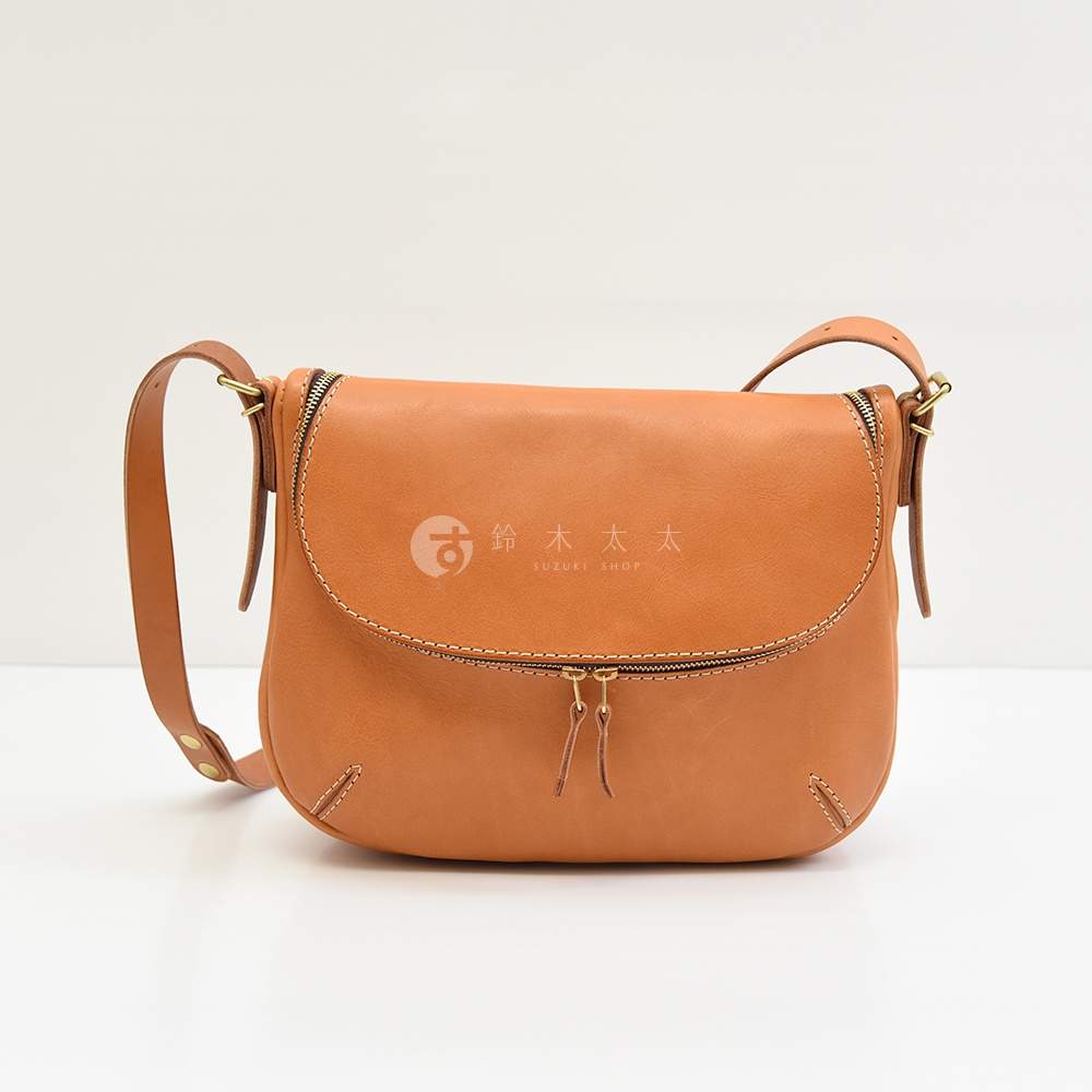 一個棕褐色的皮革馬鞍包，包身有拉鍊設計，正面有兩個小皮革拉繩裝飾，包蓋上有品牌標誌和日文文字。