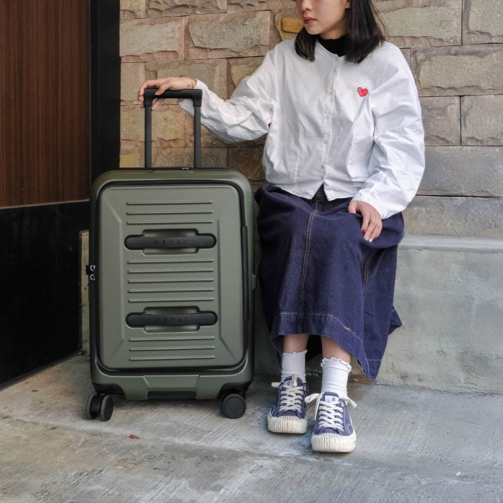 一位年輕女性坐在石牆前，手扶著一個軍綠色的 AZPAC 二十五吋前開口煞車行李箱。她身穿白色長袖襯衫、深藍色牛仔長裙與深藍色帆布鞋。行李箱表面具有橫向條紋紋理與獨特的雙手把設計，整體呈現休閒旅遊風格。