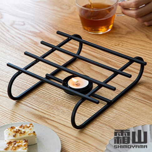 黑色的鐵藝烤漆溫茶架，結構簡潔，帶有兩個弧形支腳，架上置有一個燃燒中的茶蠟，下方有小蠟燭杯。