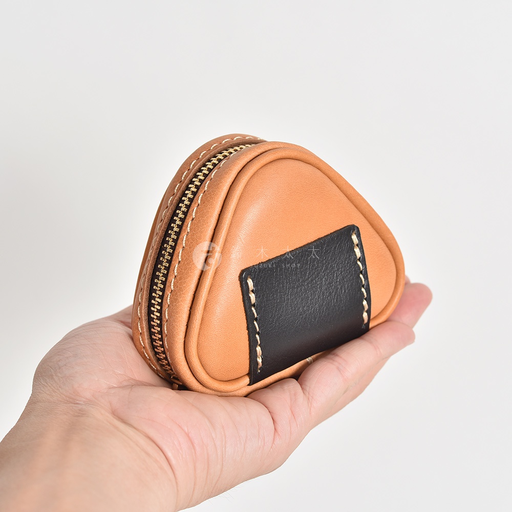 手持一個三角飯糰造型的棕色皮革小物收納包，包身為光滑皮革，飾有黑色皮革塊，以白色縫線和金色拉鍊裝飾。