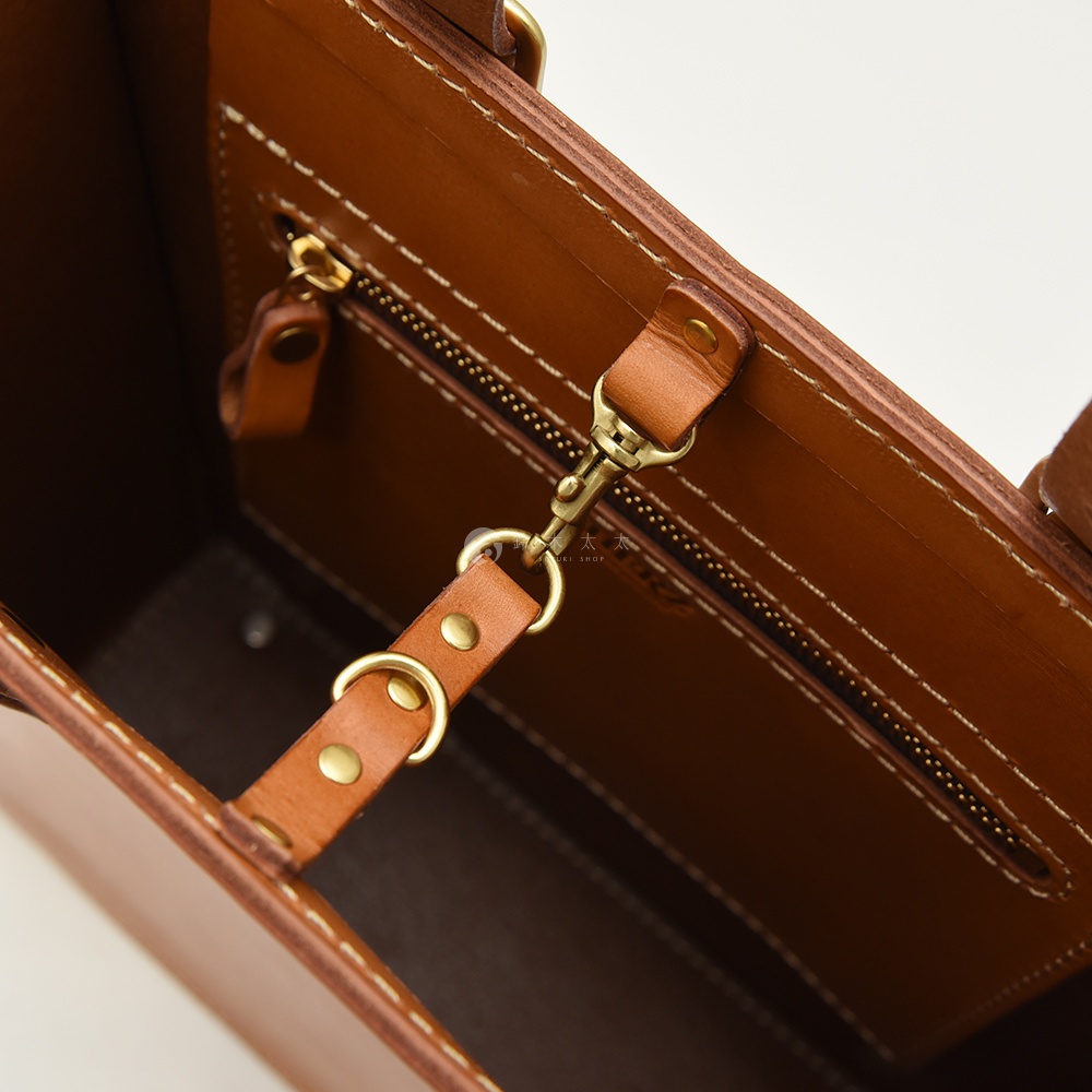 棕色皮革托特包內部細節，特寫拉鍊、皮革提帶、黃銅掛鉤及D型環，縫線清晰，展現職人手作質感。
