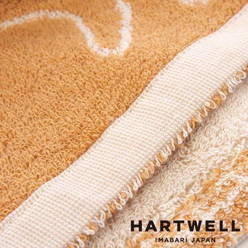 一張特寫視角的 Hartwell 今治毛巾照片，展現柔軟厚實的織物紋理，毛巾主體為溫暖的焦糖棕色，邊緣處呈現白色織邊與部分米白色毛巾細節，底部角落標註有 Hartwell Imabari Japan 品牌字樣。