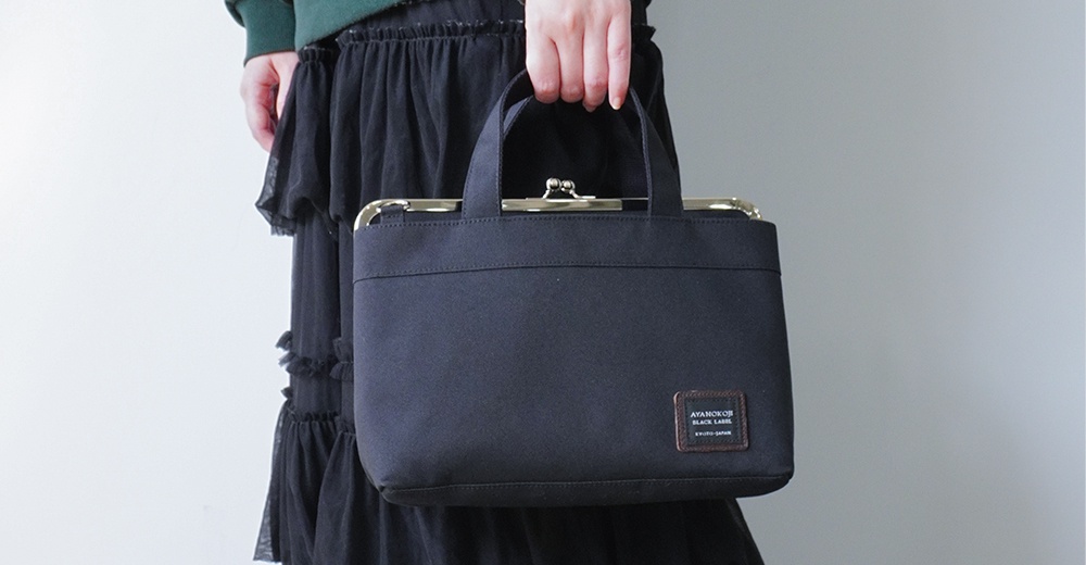 一名女子手提一個黑色方形手提包，包上有一個金屬扣，包的下方有一個小標籤，上面寫著 AYANOKOJI BLACK LABEL KYOTO-JAPAN。