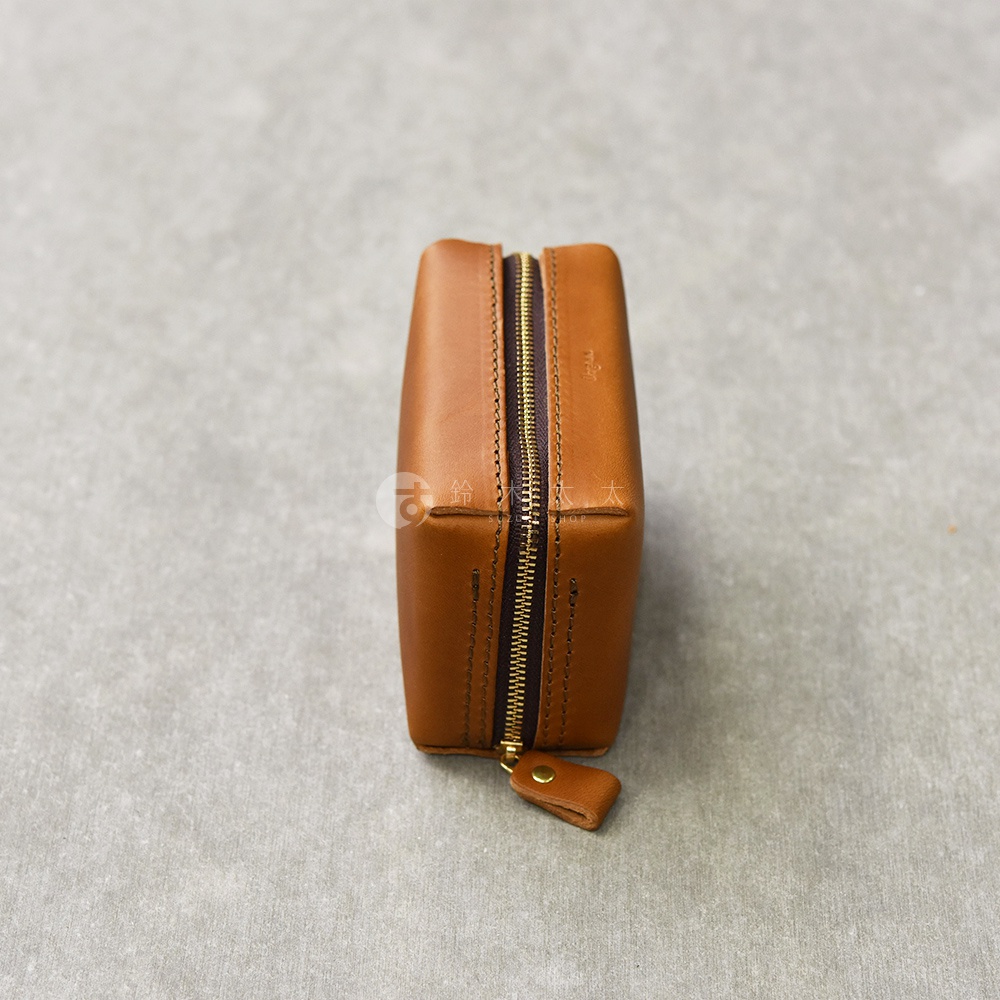 一個方形的咖啡色皮革收納包，正面有金屬拉鍊，拉鍊頭處有一個小皮片，包包擺放在灰色紋理的背景上。