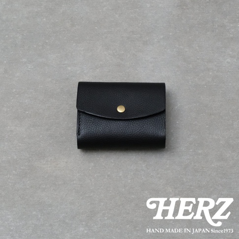 這款日本HERZ職人手作黑色皮革迷你短夾，採用質感細膩的Minerva Box皮革製成，正面中央配有黃銅色金屬按扣，設計簡約洗鍊，右下角印有白色HERZ品牌標誌，整體呈現深沉雅緻的手工藝質感。