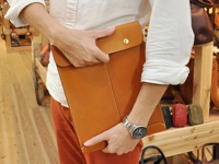 職人手作皮革信封式雙釦手拿袋，採用棕褐色光滑皮革製成，設有金色雙釦設計，車縫線清晰，整體呈現簡約風格。