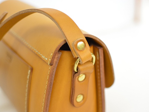 職人手作皮革寬扣帶馬鞍包，採用天然鞣製皮革，呈現溫潤的焦糖色調，搭配黃銅色的金屬扣件，展現簡約復古風格。包身線條俐落，縫線精緻。