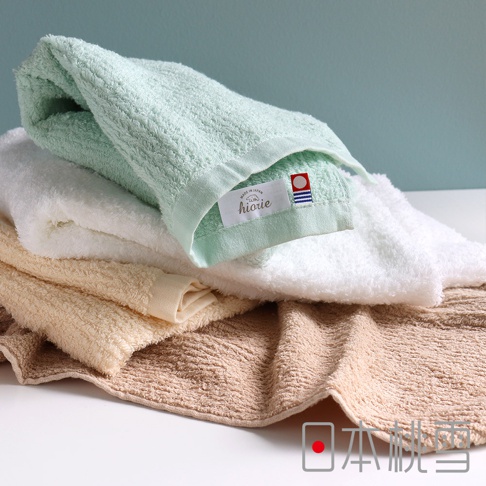 疊放的今治細絨浴巾，包含淺綠色、白色、米色和褐色。淺綠色浴巾的邊緣有一個標籤，上面印有「hiorie」字樣和「MADE IN JAPAN」的字樣。