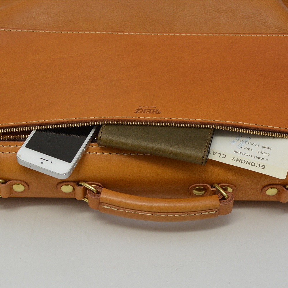 職人手作皮革旅行商務兩用波士頓包，呈現自然棕色，包身結構由皮革製成，拉鍊處可見一支白色手機、一個綠色卡夾以及一張寫有「ECONOMY CLASS」的機票。