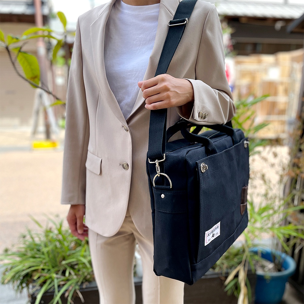 身著米色西裝外套的人，肩背一個黑色純綿帆布公事包，包款可肩背或手提，側面有品牌標籤。