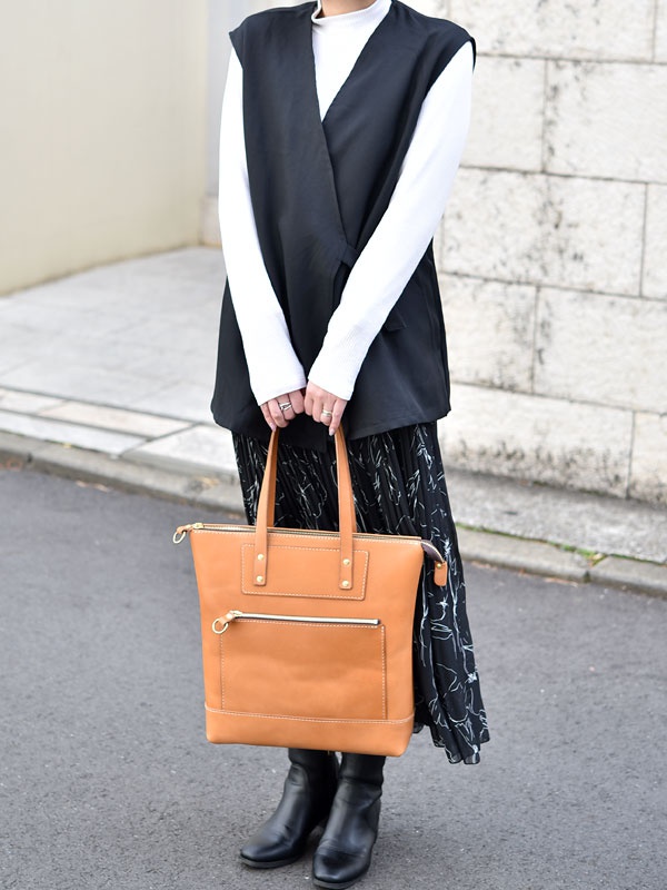 棕褐色皮革直式拉鍊托特包，正面設有拉鍊口袋及兩個金屬釘裝飾，包身縫線清晰，搭配金色拉鍊。