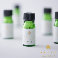 一瓶綠色玻璃瓶裝的佐賀楠木精油，瓶身上有白色標籤，印有金色「WACCA essential oil」字樣和圖案。瓶蓋為黑色。背景有幾瓶相同的精油瓶，呈現模糊的綠色。