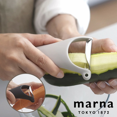 一隻手正使用灰色 Marna 品牌削皮器削去小黃瓜的外皮，削出的薄片落在下方的碗中，畫面左下角的小圓框則展示了使用黑色同款削皮器削紅蘿蔔的情境，整體呈現簡約的生活美學質感。