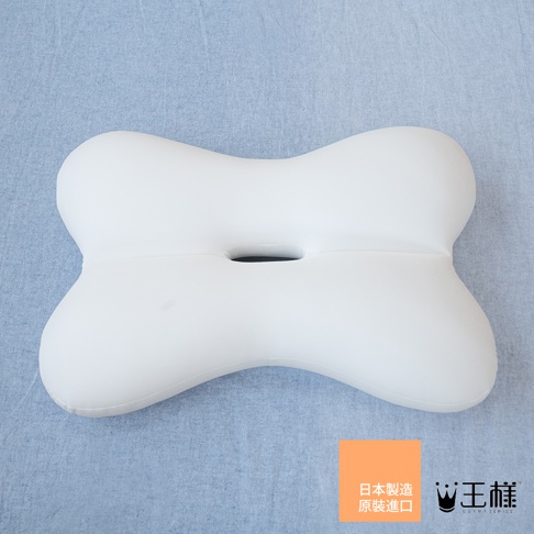 這是一款王樣品牌製造的白色長方形枕頭，外型呈現獨特的骨頭狀弧線設計，中央設有一個凹槽洞口。枕頭放置在淺藍色背景布料上，右下角配有橘色標籤，標籤上印有日文字樣及王樣品牌名稱。