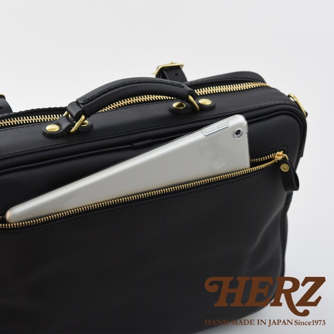 黑色皮革製成的手提公事包，設有金色拉鍊和提把，並有一個外側拉鍊口袋，其中插入一個銀色平板電腦。