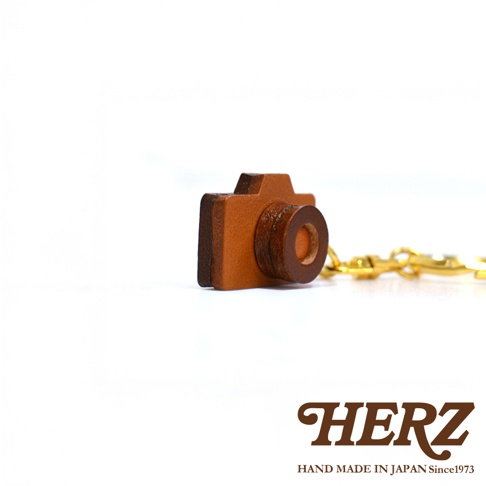 職人手作皮革相機造型鑰匙圈，主體為棕色皮革製成，造型小巧可愛，模仿復古相機，搭配金色金屬掛環。