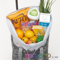 灰色印花提袋購物車內裝有數樣商品，包括橘子、檸檬、番茄、青椒、洋芋片、優格、牛奶盒、麵食包以及一小叢綠色植物。