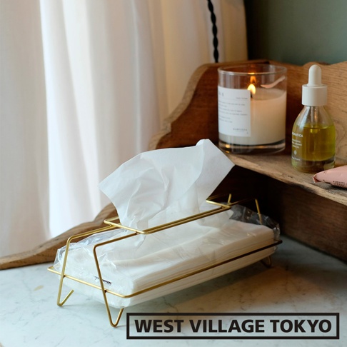 黃銅色金屬支架，內置白色面紙盒，裝有整齊堆疊的白色面紙，面紙盒置於大理石紋桌面，背景為白色窗簾、燃燒的蠟燭和一瓶精油。