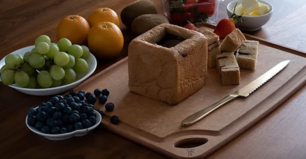 金屬麵包刀，刀刃呈鋸齒狀，附有金屬握柄，放置於木製砧板上，旁邊有切塊的麵包、藍莓、綠葡萄、柳橙、奇異果及草莓。