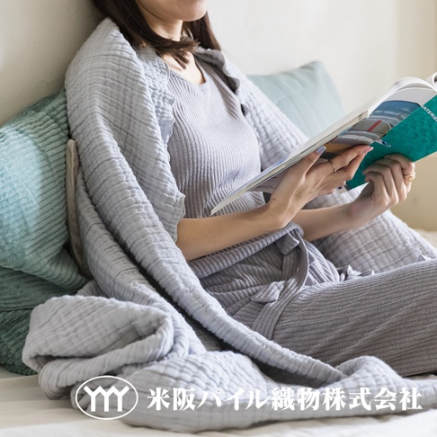 淺灰色三層紗輕透波紋四季毯，質地柔軟，輕盈透氣，覆蓋在一位正在閱讀的女性身上，她身穿同色系居家服，背景為淺藍色燈芯絨枕頭。