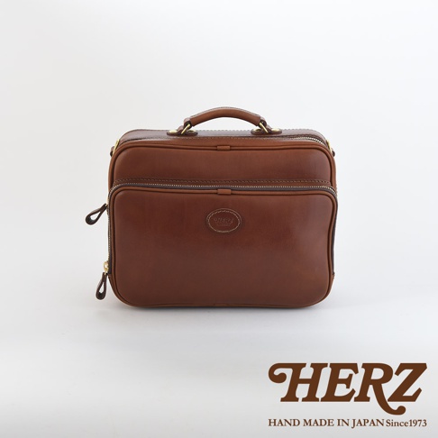 咖啡色皮革製成的手提公事包，中間有圓形品牌標誌，雙拉鍊設計，頂部有提把，兩側有可拆卸的肩帶固定點。