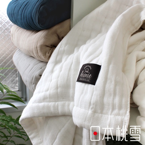 白色八層紗透氣被，材質為棉，柔軟親膚，帶有品牌標籤。旁邊堆疊著深藍、米色和灰色等不同顏色的被子。