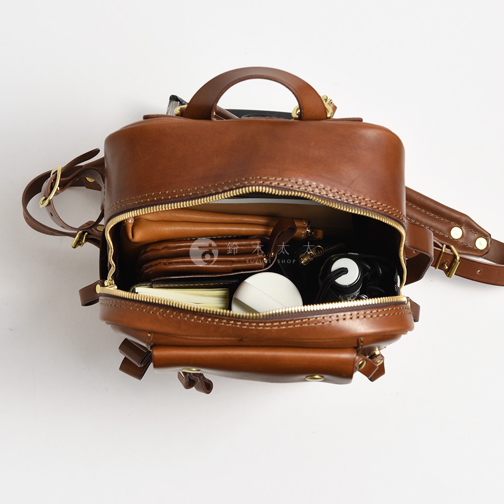 職人手作皮革兩用多功能後背包 R-66，展示棕色皮革材質，包款敞開，內部可見皮夾、筆記本、白色圓形物品與黑色線狀物品，搭配金色拉鍊與金屬扣環。