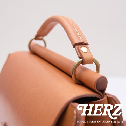 HERZ 職人手作皮革屋瓦風兩用單肩包，近距離拍攝其皮革提把與金屬環細節，並顯示品牌標誌。
