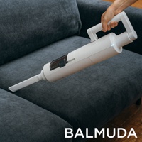 白色The Cleaner無線吸塵器，具有可調節提把，正被手持清潔深灰色布藝沙發。吸塵器主體為圓筒形，配有透明集塵盒和扁平吸嘴。