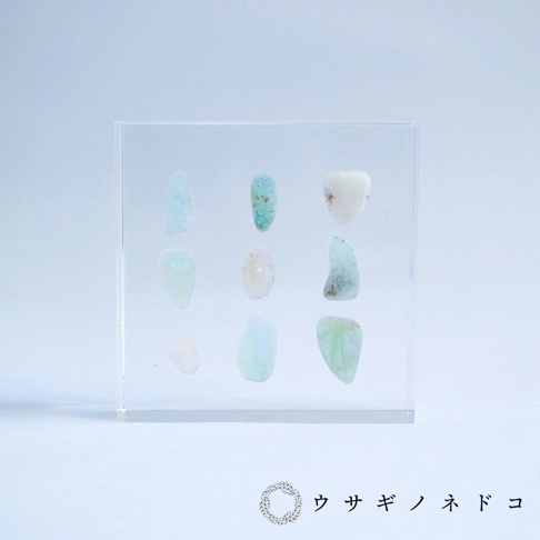 透明方形壓克力塊中，以九宮格方式陳列著九顆近似橢圓形的細石標本，顏色包含淺藍、淺綠、淡粉及白色，部分標本帶有天然紋理。