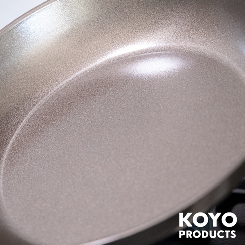 從斜上方近距離拍攝的KOYO PRODUCTS輕量壓紋淺型鐵製平底鍋，鍋身呈現帶有細緻金屬光澤的淺褐色，內表面分布著均勻的壓紋質感，右下方印有白色品牌字樣。