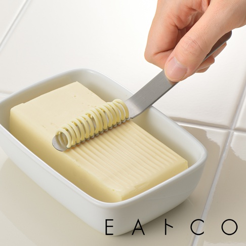 日製不鏽鋼奶油刀，刀刃處有螺旋狀切槽，正用於刨出奶油條，置於白色奶油盒中。