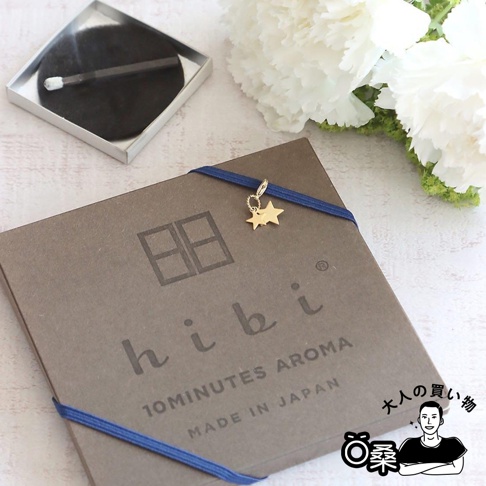 灰褐色紙盒裝有三入組香氛火柴，盒面印有「hibi 10MINUTES AROMA MADE IN JAPAN」字樣，並以藍色緞帶綁住，緞帶上掛有金色雙星吊飾。左上角為一個銀色方形盒，內有燃燒中的香氛火柴，背景為白色花朵。