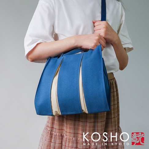 藍色棉帆布扇形托特包，搭配米白色帆布拼接，側面有兩條放射狀的縫線。包包由一人提著，肩背一條同色系的肩帶。