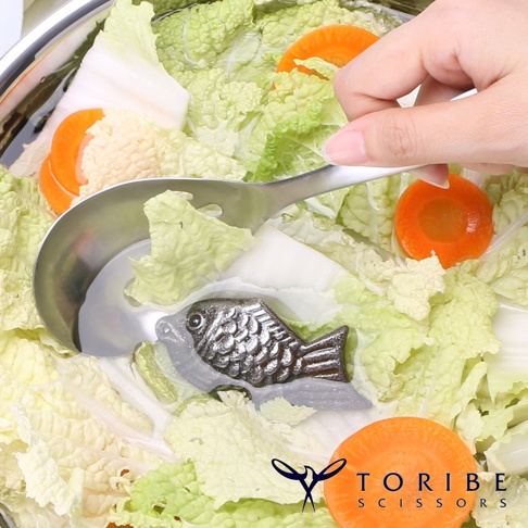 鍋中盛有白菜、紅蘿蔔等蔬菜，一把不鏽鋼湯勺正撈起蔬菜，湯勺底部有一個銀色、造型為小鯛魚的補鐵塊。