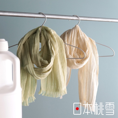 兩條皺褶質感的純棉紗布圍巾懸掛在銀色金屬衣架上，左側為淡綠色，右側為米白色，背景為淡藍色牆面，前景左下方可見白色瓶罐，右下角印有日本桃雪品牌標誌。
