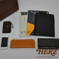 皮革製品組合，包含一個深棕色大皮夾、一個綠色卡夾、一本黑色筆記本、兩本雜誌、一個橘色長皮夾、一個黑色長皮夾，以及一支白色手機。