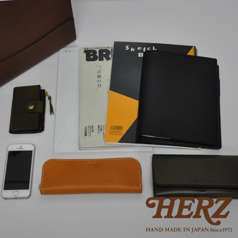 皮革製品組合，包含一個深棕色大皮夾、一個綠色卡夾、一本黑色筆記本、兩本雜誌、一個橘色長皮夾、一個黑色長皮夾，以及一支白色手機。