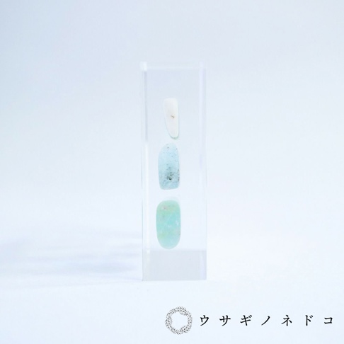 透明壓克力塊中垂直排列三顆片狀標本，由上至下依序為白色、淺藍色和綠松石色，呈現天然石材紋理。