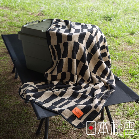 日本桃雪今治戶外格紋棉柔浴巾，擺放在露營椅上，浴巾上有黑米格紋，一角有Hiorie品牌橘色標籤。