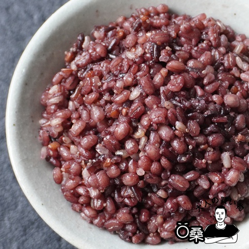 一碗裝滿紅褐色十六穀米的特寫，穀物飽滿，質地濕潤，碗邊為淺灰色粗陶材質。