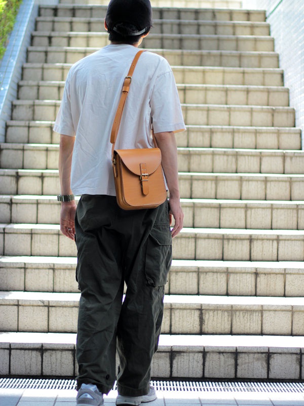一位男士斜挎著一個棕褐色皮革翻蓋迷你單肩包，包包上有金屬扣環和可調節肩帶。