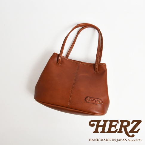 HERZ 品牌標誌性的棕色皮革托特包，帶有可調式提把，包身上壓印有 HERZ 品牌字樣，整體採用簡約設計，呈現職人手作質感。