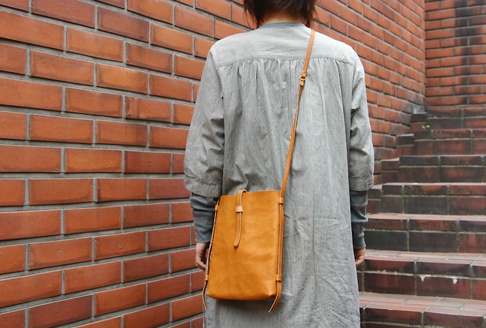 咖啡色皮革製直式單肩包，配有同色系細長肩帶，包身設計簡約，以皮扣固定開口。