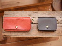 兩個職人手作皮革收納包，一個為紅棕色，另一個為深棕色，皆為長方形，帶有金色圓形按扣和金屬環，置於木質表面上。