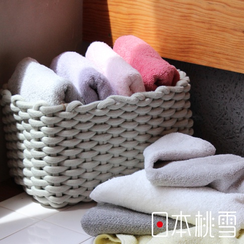 一籃淺灰色編織毛巾，內有粉色、紫色、淺紫和白色毛巾。籃子旁邊堆疊著白色、淺灰、深灰色和淺黃色毛巾，材質柔軟。